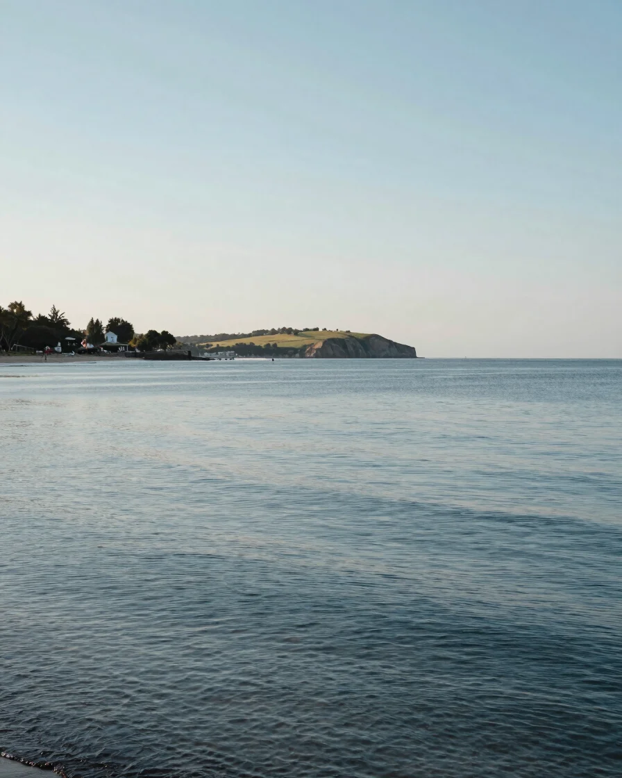 Wide environmental view of a coastal shoreline at midday in soft natural daylight, calm water and distant headland, true
